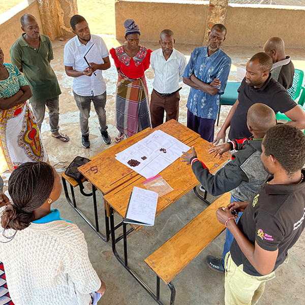 group of people standing around a table, listening to a speaker