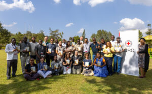 A group shot of all of the SEADS training participants in the sunshine holding their SEADS Handbooks and standing next to an ICRC banner